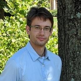 Man with short dark hair wearing glasses and blue button up shirt standing in front of a tree