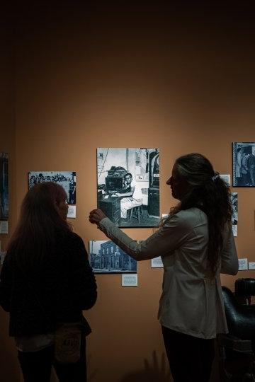 Seen from behind, two people look closely at black-and-white photos displayed on an orange wall. 